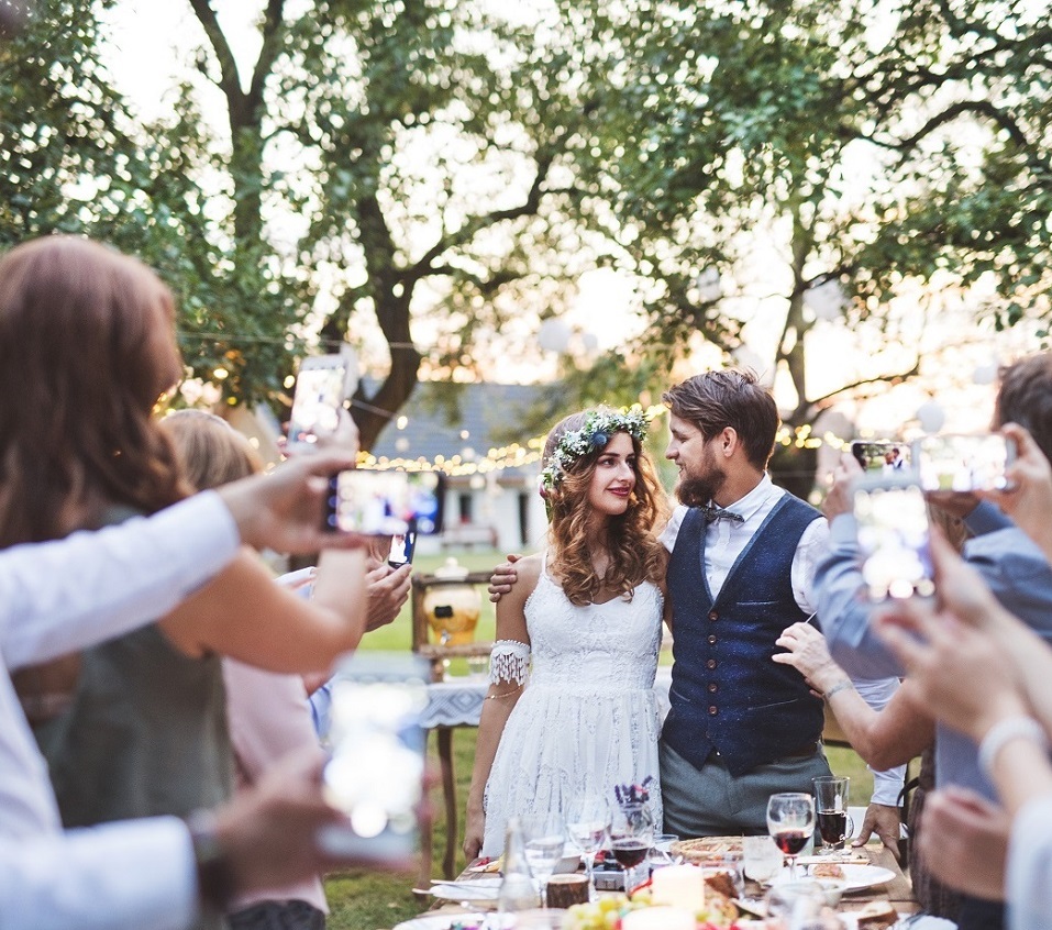 Guests with smartphones taking photo of bride and groom at wedding reception outside in the backyard.